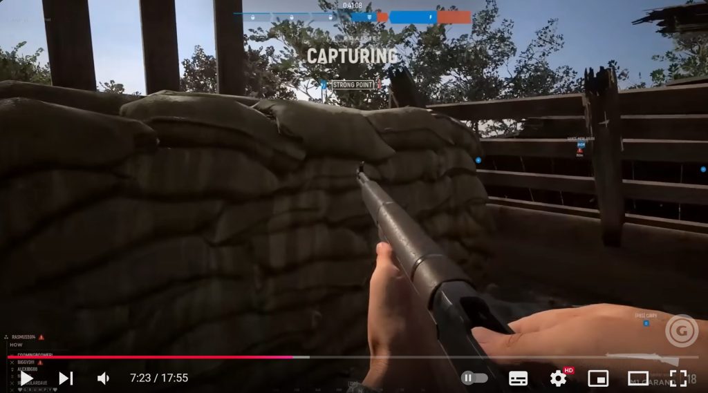 An image from a first-person shooter game, inside a trench, with sandbags ahead, wooden fencing to the right, and a gun held in both hands in the centre of the image.