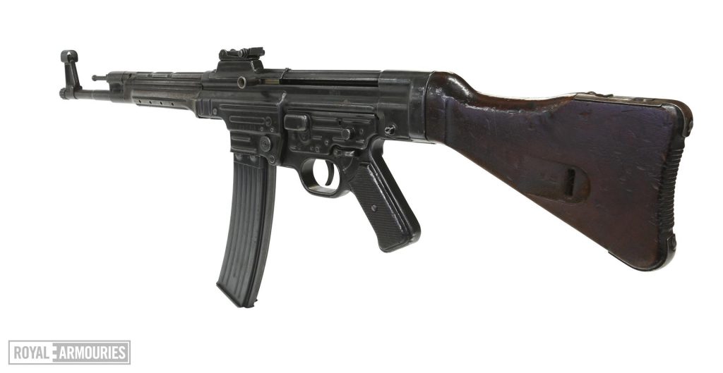 An assault rifle, pointed to the left. The gun is black, with a long magazine curving forwards and a wooden stock.