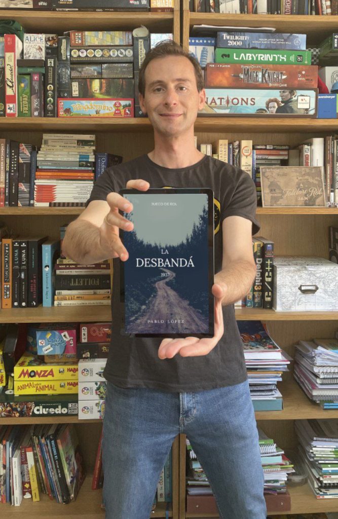 A white man with short brown hair and dark eyes wearing a brown t-shirt and blue jeans, standing in front of book shelves holding board games and books. He holds a tablet out in front of him, displaying the cover of the digital copy of the La Desbandä 1937 roleplaying game.